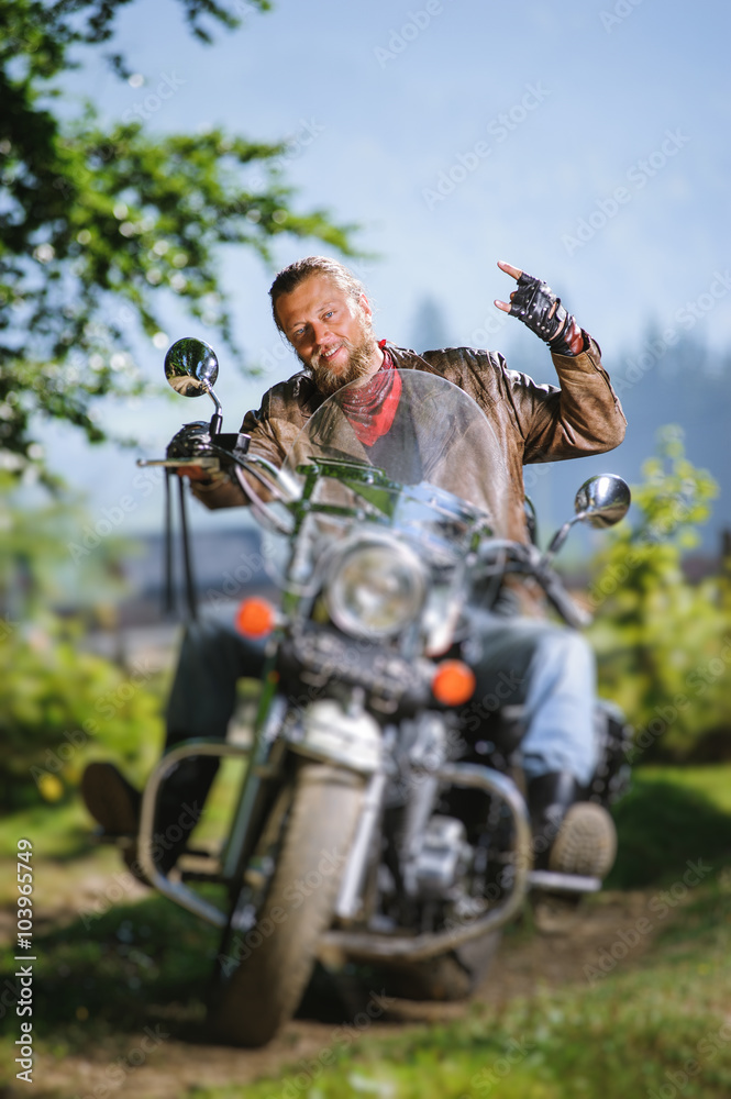 Handsome biker with beard driving his cruiser motorcycle in the forest and giving the devil horns gesture and smilling. Man is wearing leather jacket and blue jeans. Tilt shift lens blur effect