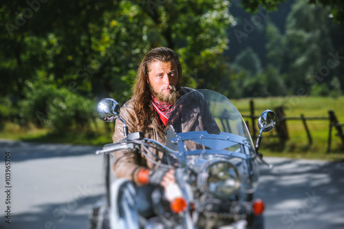 Portrait of biker with long hair and beard in a leather jacket and sunglasses sitting on his bike beside the road in the forest. Looking into the camera. Tilt shift lens blur effect photo