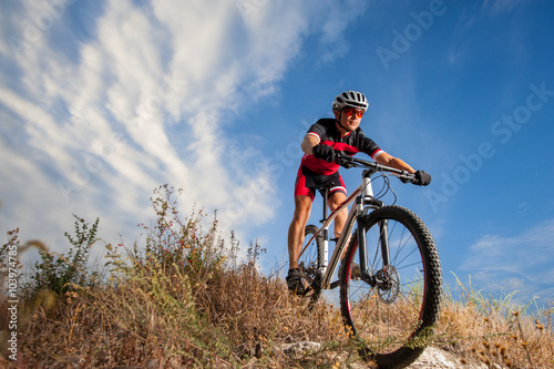 Cyclist riding his bike down on mountain trail. Biker is wearing red sportswear helmet and red glasses. Low point of view. Beautiful sky and clouds on backgraund