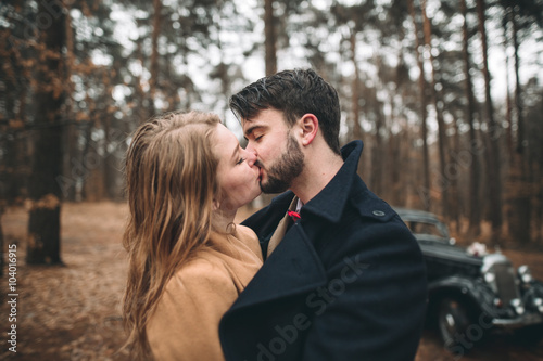 Romantic fairytale wedding couple kissing and embracing in pine forest near retro car. © olegparylyak