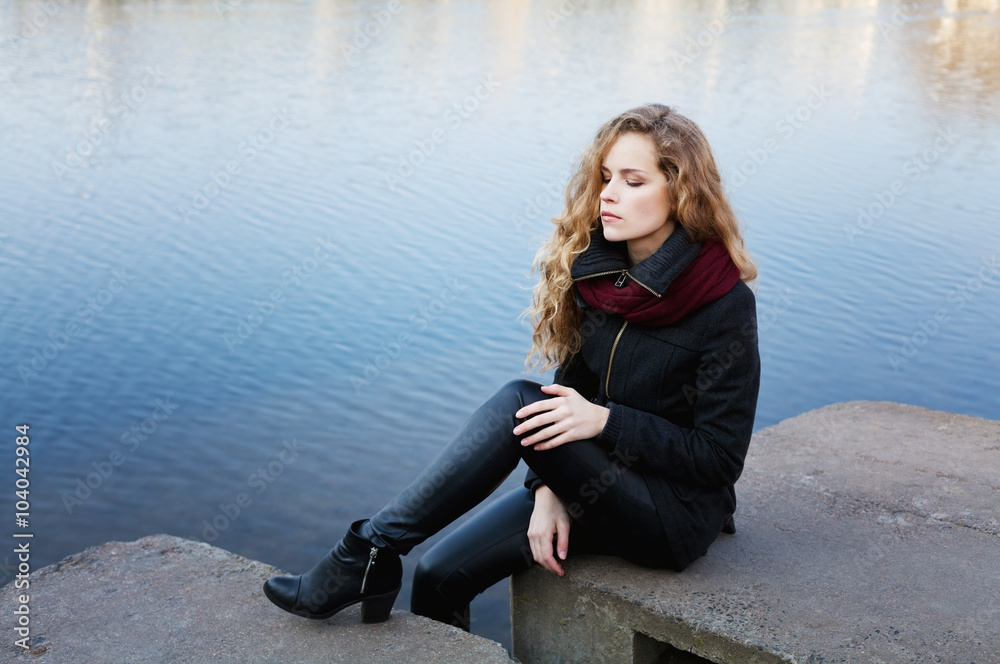 beautiful blonde woman with long curly hair sitting on the banks of the blue river water having closed eyes and enjoying silence, meditation or relaxation concept