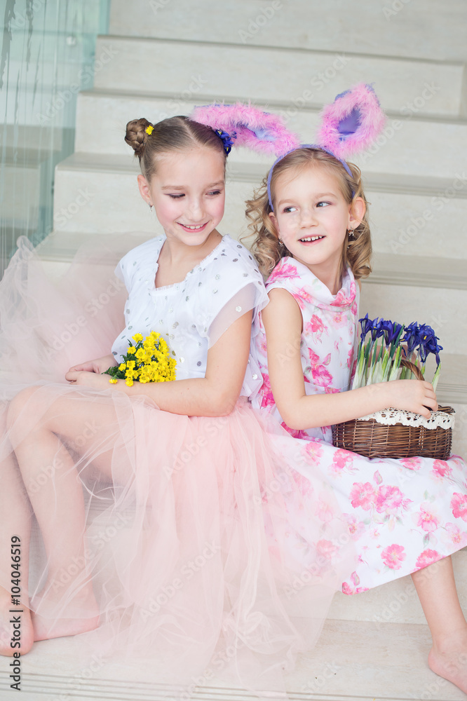 Two cute sisters sitting indoors with bunny ears and flowers celebrate Easter and spring Two cute sisters sitting indoors with bunny ears and flowers celebrate Easter and spring