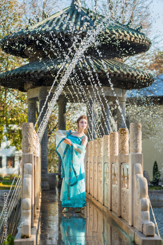 pretty young geisha in water drops in blue dress standing near fountain