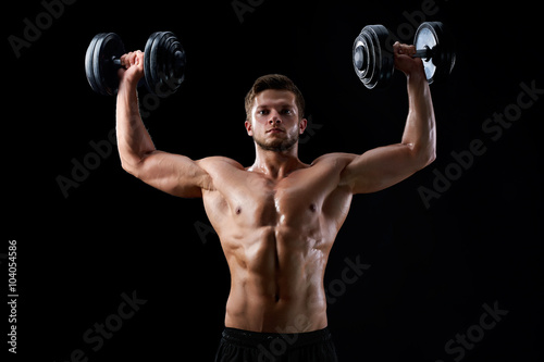 Reaching out for goals. Young serious muscular handsome man holding up two dumbbells exercising on black background