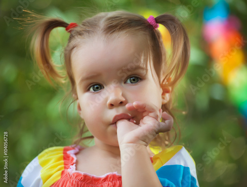 cute little girl wearing colorful dress sucks her thumb. Bad habit. in the park