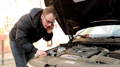 Young, handsome man in black jacket and glasses standing behind his car and speaking thru the phone probably about his engine. He has problem because he doesn’t know what to do.