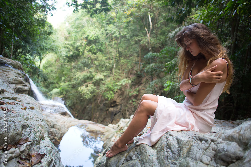 The beautiful fashionable girl with long hair, sits on surface stones, dressed in a light white dress, finding on tropical falls of the island Samui. Sunny day and happy smile. The beautiful fashionable girl with long hair, sits on surface stones, dressed in a light white dress, finding on tropical falls of the island Samui. Sunny day and happy smile.