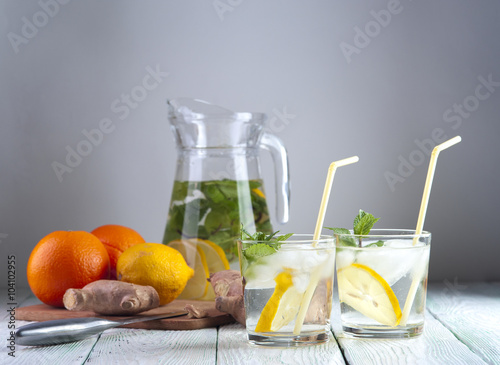 Glasses of detox drink with mineral water, lemon and mint, glass jug, lemon, orange and ginger on wooden table. Drink for diet. Focus on glasses.