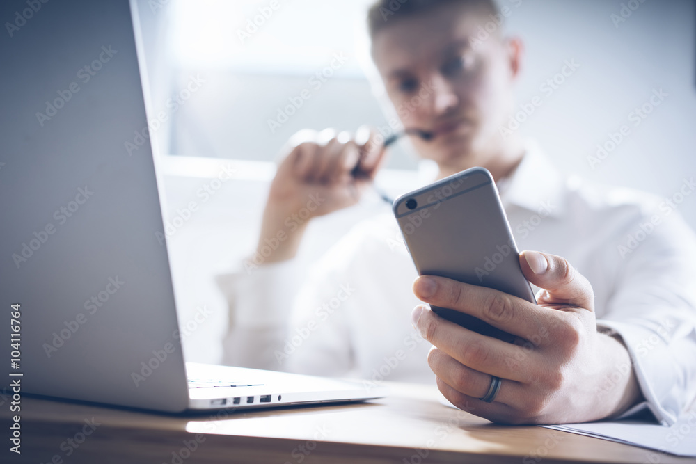 Close up image of serious businessman using smart phone and laptop at his office, successful man employer sitting with cellphone in office interior and looking concentrated Close up image of serious businessman using smart phone and laptop at his office, successful man employer sitting with cellphone in office interior and looking concentrated