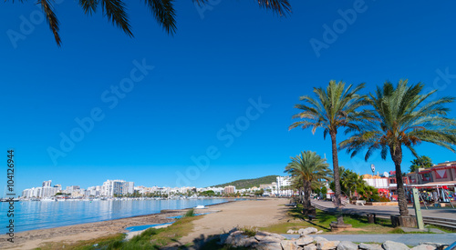 Mid morning sun, walk along beach near the city. Rows of palm trees line the beach, sunny day along water's edge in Ibiza, St Antoni de Portmany Balearic Islands, Spain.