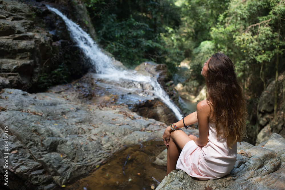 The beautiful fashionable girl with long hair, sits on surface stones, dressed in a light white dress, finding on tropical falls of the island Samui. Sunny day and happy smile. The beautiful fashionable girl with long hair, sits on surface stones, dressed in a light white dress, finding on tropical falls of the island Samui. Sunny day and happy smile.