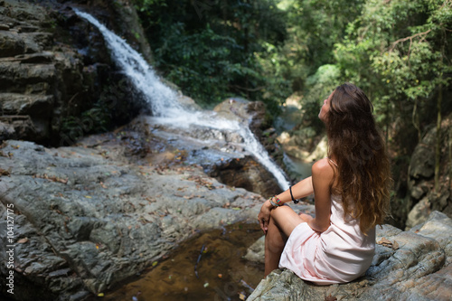 Fototapeta Naklejka Na Ścianę i Meble -  The beautiful fashionable girl with long hair, sits on surface stones, dressed in a light white dress, finding on tropical falls of the island Samui. Sunny day and happy smile. 