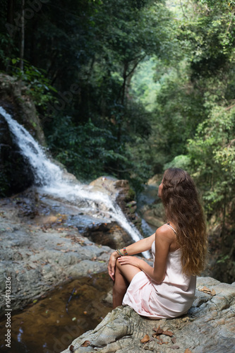 The beautiful fashionable girl with long hair, sits on surface stones, dressed in a light white dress, finding on tropical falls of the island Samui. Sunny day and happy smile.