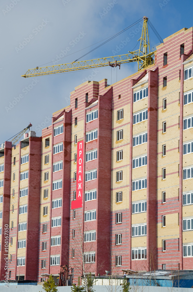 Construction crane and housing under construction./ New modern apartment houses in the course of construction. On the house the poster with an inscription in Russian - sale.