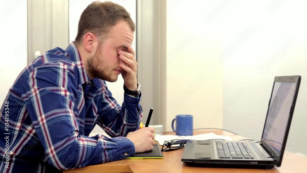 Young men in colrfull shirt, sitting behind his desk and doing something on his computer, he is tired and mad because of his work to do. Young men in colrfull shirt, sitting behind his desk and doing something on his computer, he is tired and mad because of his work to do.