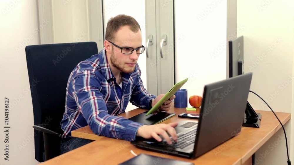Good looking men sitting behind his desk, using tablet and talking thru the phone. He is writing something into his note. Good looking men sitting behind his desk, using tablet and talking thru the phone. He is writing something into his note.