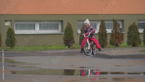 Kid Tries to Sit on a Bike Wheeling Little Girl is Riding a Two Wheeled Bicycle by a School Yard Lawn Evergreen Trees Wet Road Puddles Winter or Fall
