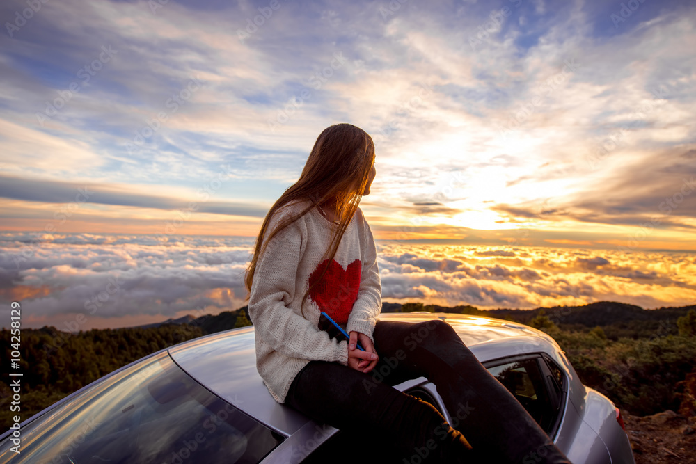 Fototapeta premium Young woman in sweater with heart shape enjoying beautiful cloudscape sitting on the car roof above the clouds on the sunrise