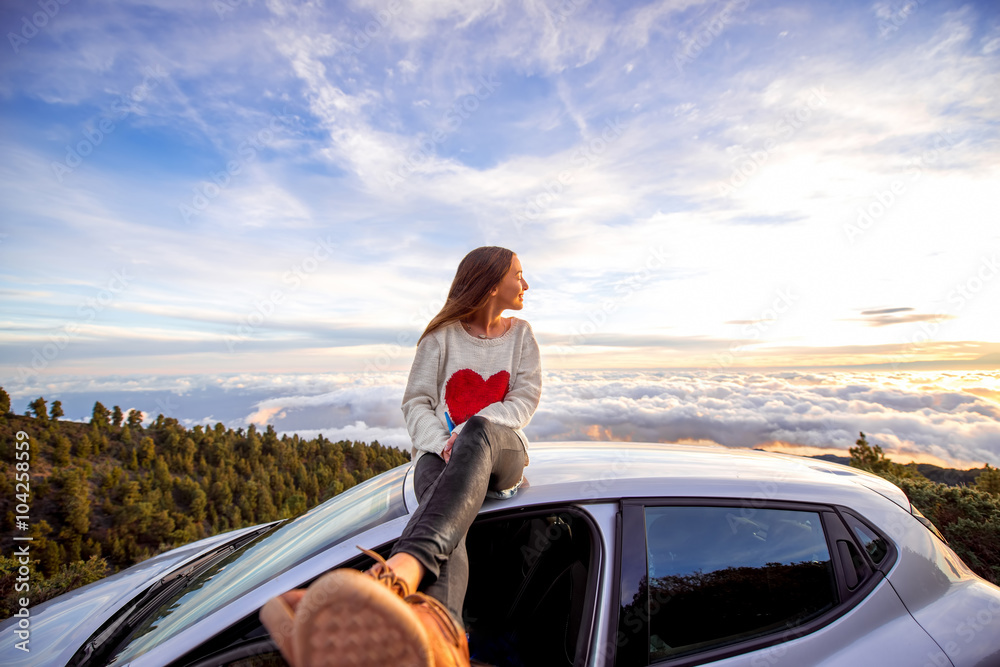Young woman in sweater with heart shape enjoying beautiful cloudscape sitting on the car roof above the clouds on the sunrise Young woman in sweater with heart shape enjoying beautiful cloudscape sitting on the car roof above the clouds on the sunrise