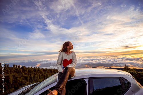 Young woman in sweater with heart shape enjoying beautiful cloudscape sitting on the car roof above the clouds on the sunrise © rh2010