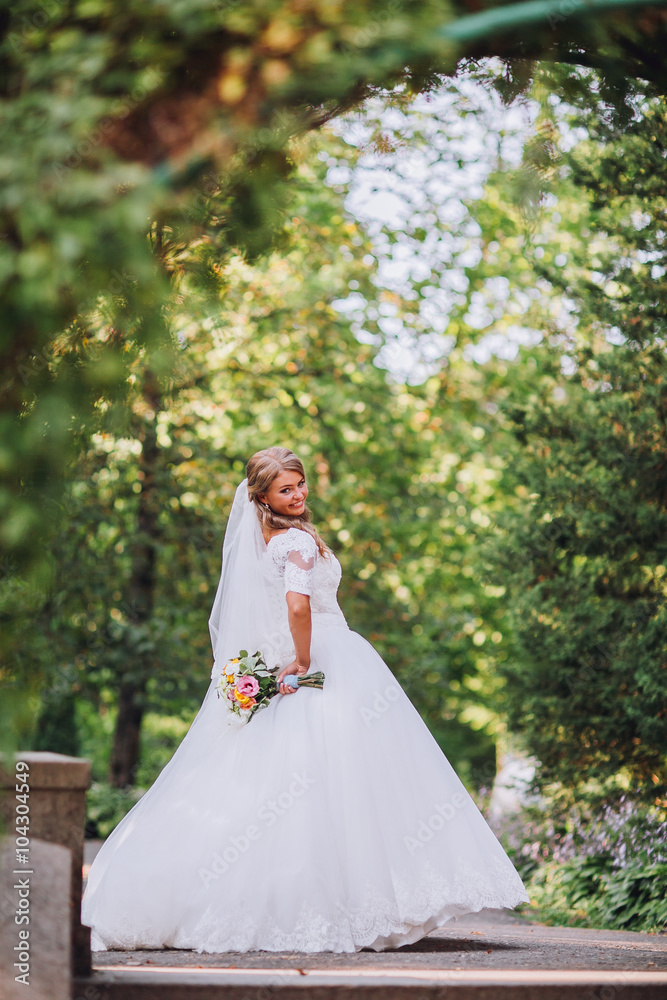 Gorgeous blonde bride in elegant dress holding bouquet and posing in the sunny summer park or garden on their wedding day