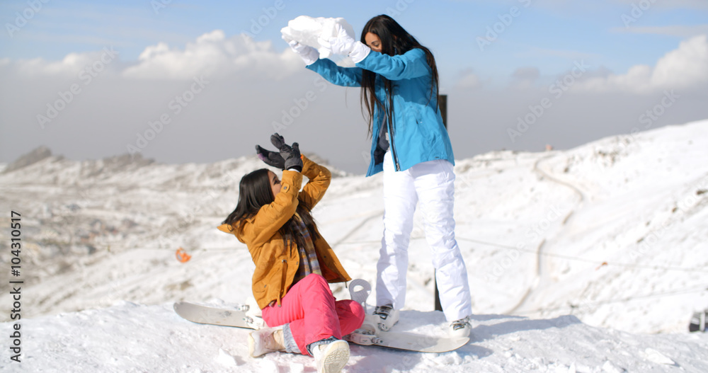 Two young female friends playing in the snow threatening to cover one with a large snow ball as she sits on her snowboard in the snow on a mountain summit.
