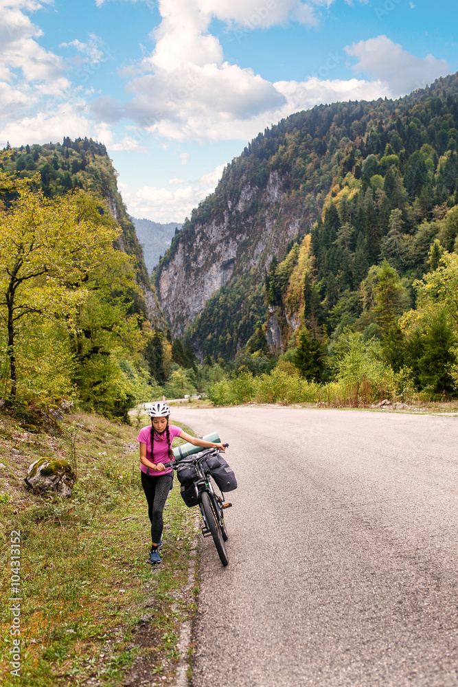 Young woman with backpack and bicycle walking on countryside terrain. Tourist emerging from a lengthy uphill, mountains in the background