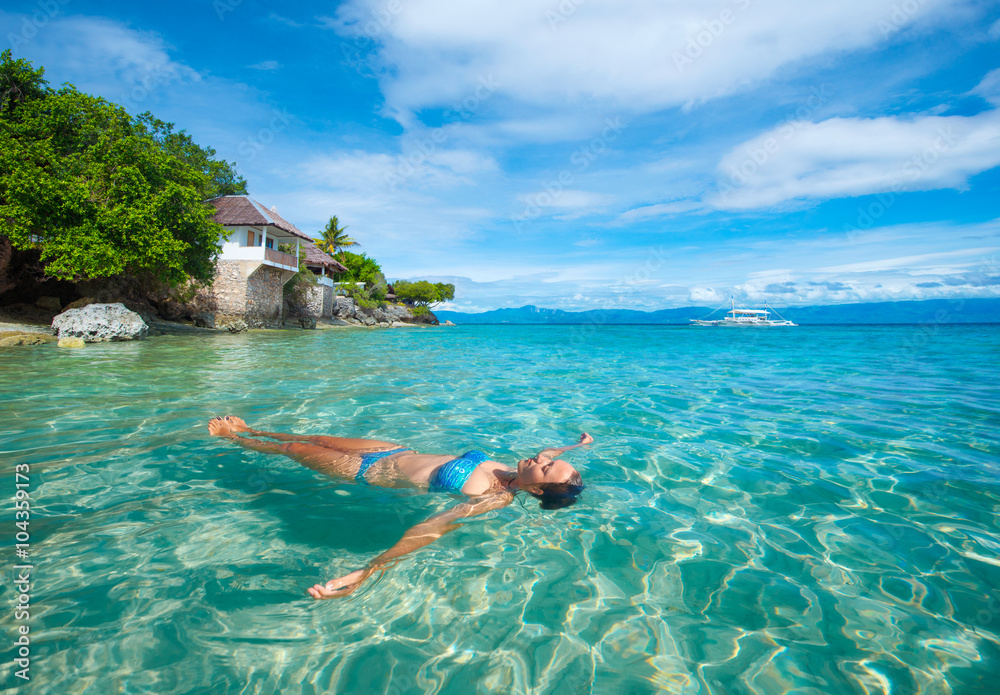 young woman in bikini relaxing lying on water against background beach and bungalow