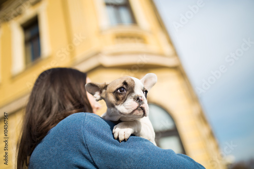 Young casually dressed woman holding her adorable French bulldog puppy. Close up shot with wide angle lens. Old, rustic building in background. © Dusko