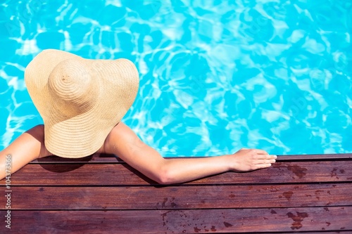 Obraz na płótnie Woman wearing straw hat leaning on wooden deck by poolside