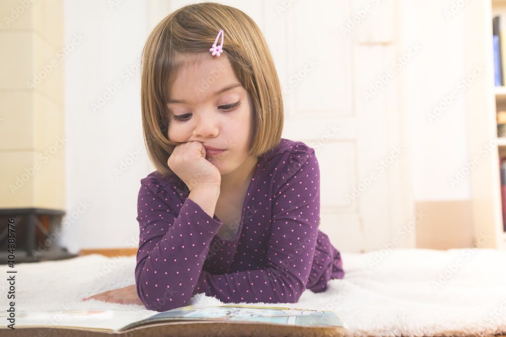 Pensive little girl lying on blanket on the floor watching picture book Pensive little girl lying on blanket on the floor watching picture book