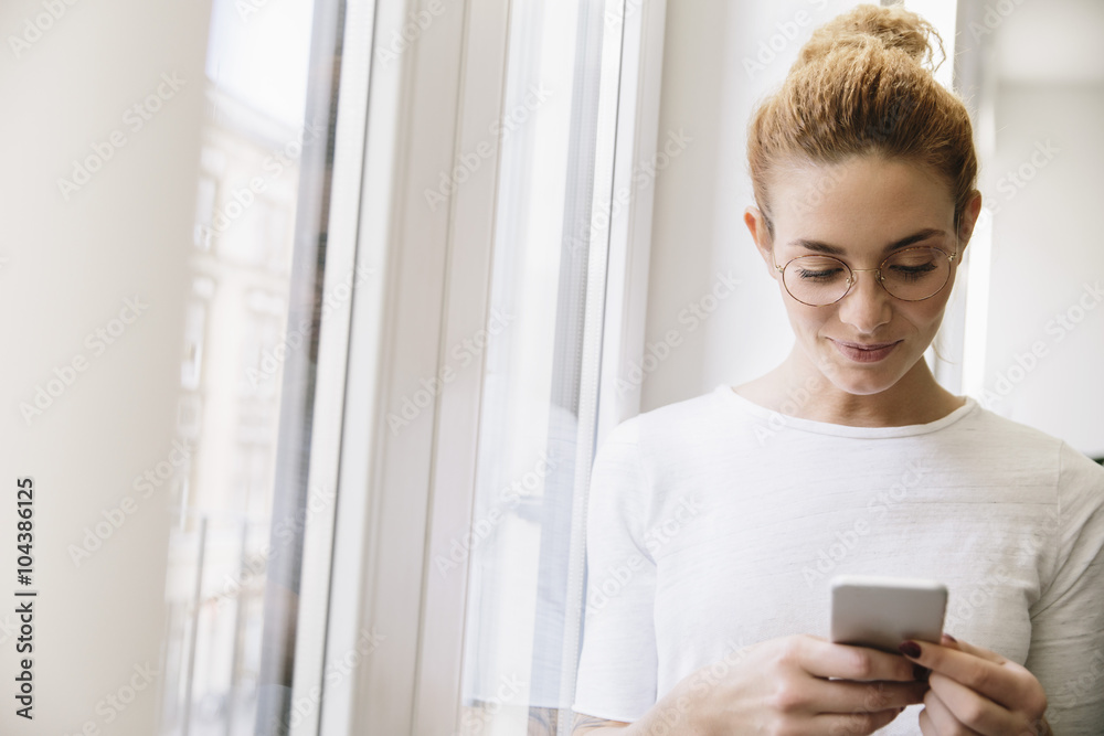 Young woman looking at her mobile phone, standing by window Young woman looking at her mobile phone, standing by window