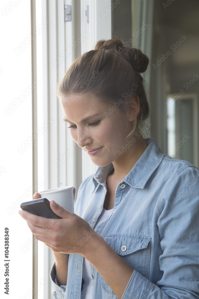 Young woman standing at the window with smart phone and coffee cup Young woman standing at the window with smart phone and coffee cup