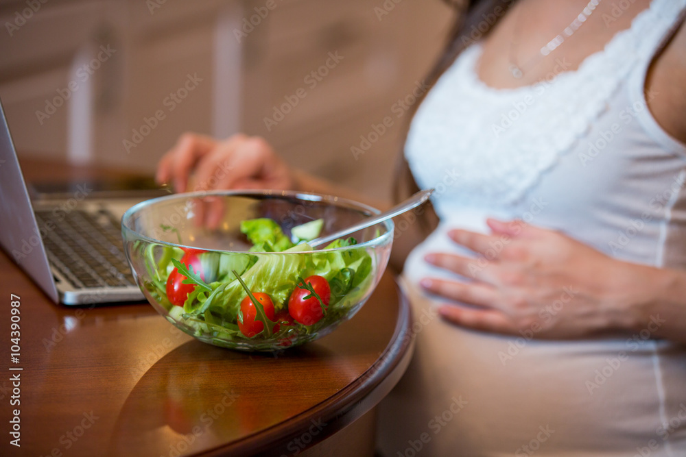 A young beautiful pregnant woman eating a fresh green vegetable salad at the table. Lady having lunch in the kitchen and using the laptop. Healthy nutrition and pregnancy. Healthy lifestyle.