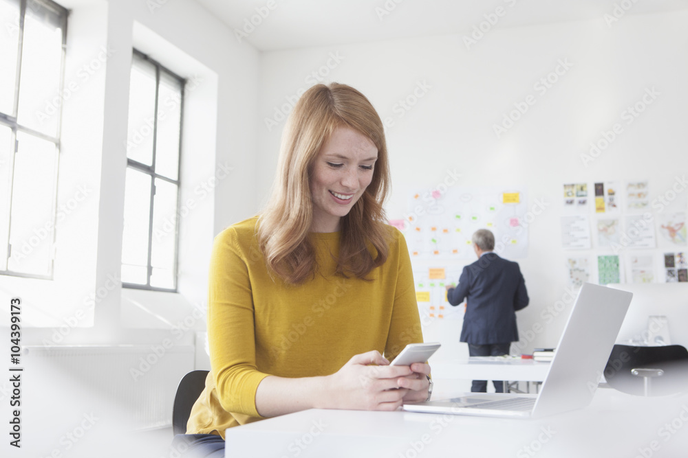 Smiling young woman in office at desk with laptop and cell phone Smiling young woman in office at desk with laptop and cell phone