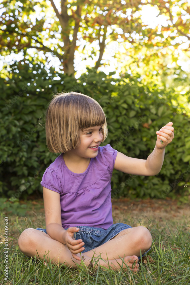 Smiling little girl sitting on a meadow watching something in her hand Smiling little girl sitting on a meadow watching something in her hand