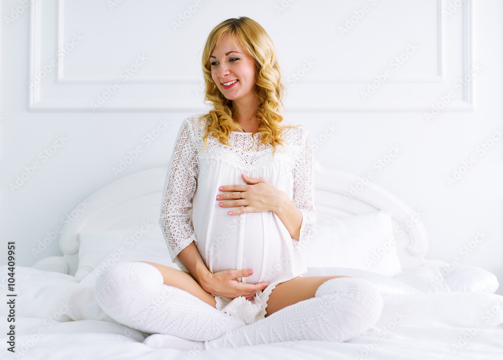 The pregnant woman sits on a bed in white bedroom The pregnant woman sits on a bed in white bedroom