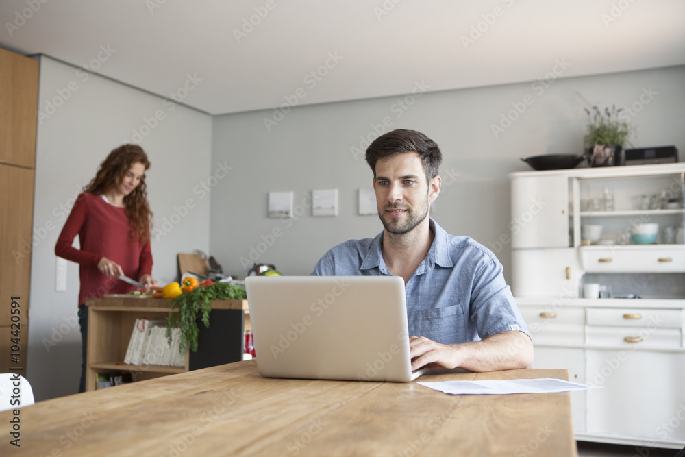 Man at home using laptop with woman in background cutting vegetables Man at home using laptop with woman in background cutting vegetables