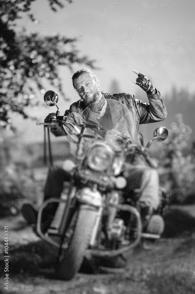 Naklejka premium Handsome biker with beard driving his cruiser motorcycle in the forest and giving the devil horns gesture and smilling. Man is wearing leather jacket and jeans. Tilt lens blur effect. Black and white