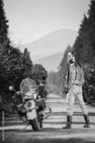Handsome biker with long hair wearing leather jacket, jeans, boots and gloves standing near his custom made cruiser motorcycle on the open road. Looking to the sun. Tilt soft effect. Black and white