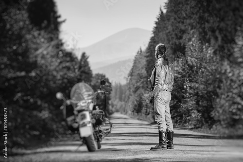 Biker with long hair wearing leather jacket jeans leather boots and gloves standing near his custom made cruiser motorcycle on the open road. Looking into distance. Tilt soft effect. Black and white