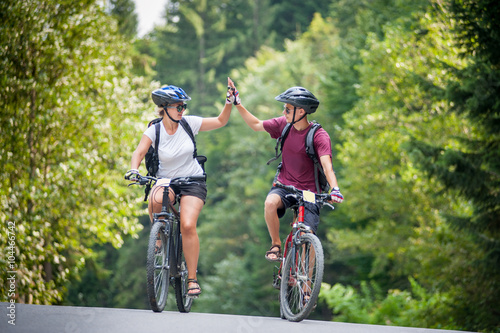 Young couple ride on a mountain asphalt road in the woods on bikes with helmets giving each other a high five