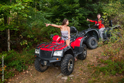 Happy beautiful couple driving four-wheelers ATV and looking at each other. Girl is pointing into the distance. Man is giving thumb up