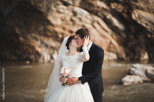 Handsome romantic groom and beautiful bride posing near river in scenic mountains