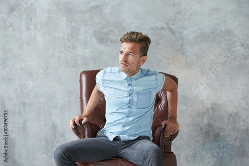 Portrait of a stylish intelligent man stares into the camera, small unshaven, charismatic, blue shirt, sitting on a brown leather chair, dialog, negotiation, short sleeve, brutal, hairstyle