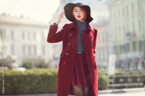 Outdoor portrait of young beautiful fashionable woman posing at street of the old city. Model wearing stylish black wide-brimmed hat, red coat. Girl looking at camera. Female fashion concept. Toned