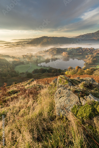 Lingering fog over Loughrigg Tarn on a fresh Autumn morning with mountain rocks in foreground.