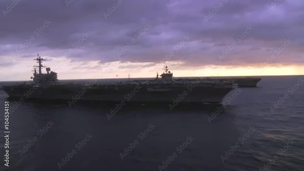 Aerial over two aircraft carriers on the high seas with helicopter moving between.