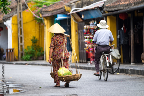 Unidentified old woman in traditional Vietnamese clothes carrying buskets with food on the street in Hoi An city. Hoian is recognized as a World Heritage Site by UNESCO.  photo