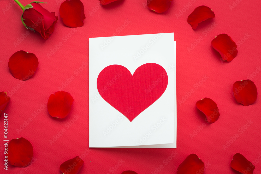 Holiday Card. Heart for Valentines day. Close up Small White Card with I Love You Message and Red Rose Flower on Top of the red background Holiday Card. Heart for Valentines day. Close up Small White Card with I Love You Message and Red Rose Flower on Top of the red background